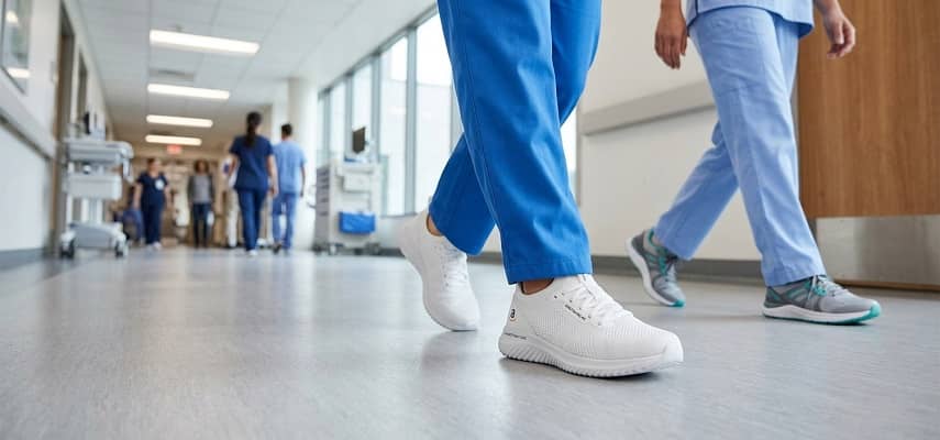 Two healthcare workers wearing blue scrubs walk down a hospital corridor; the one on the left wears white athletic shoes, and the one on the right wears gray and teal athletic shoes.