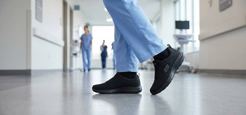 Lower legs of a medical professional wearing light blue scrubs and black slip-resistant athletic work shoes walking on a polished grey hospital corridor floor.