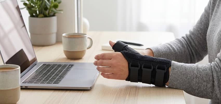 Person wearing a black thumb spica splint brace resting hand on a modern desk for joint support.
