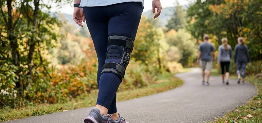 Rear view of woman in navy leggings with hinged knee brace walking on a tree-lined path.