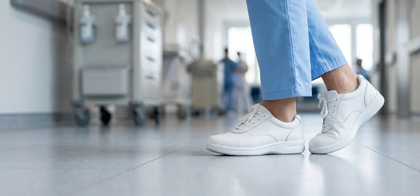 White nursing shoes and blue scrub pants are worn by a medical professional walking on a polished floor in a hospital hallway. The background shows equipment and other blurred figures.