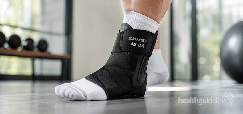 Close-up of a Zamst A2-DX ankle brace worn over a white athletic sock in a modern gym, showing supportive straps and stabilizing side panels.